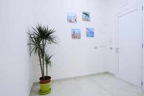 A hallway with a potted plant, four framed pictures on the wall, a white door, and a light switch. - GuesHost - Positano Apartment in Salerno (Salerno)