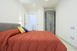 Ein Schlafzimmer mit einem großen Bett, einem grauen Kopfteil, einem Kleiderschrank und einer Tür zu einem weiteren Raum.