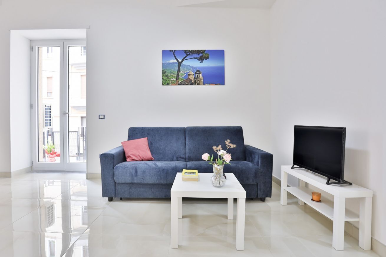 A living room with a blue sofa, a white coffee table with a vase of flowers, a flat-screen TV on a white stand, and a painting of a coastal scene on the wall.