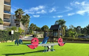Ein Spielplatz mit Wippe, Rutschen und einer kleinen Schutzhütte, umgeben von Grünflächen und einem Gebäude im Hintergrund.