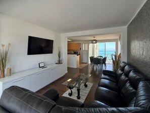 A modern living room with a black leather sofa, a flat-screen TV, a glass coffee table, and a view of the kitchen and dining area. - Costa Dorada Apartment in Salou - Beach & Wi-Fi Included (Salou)