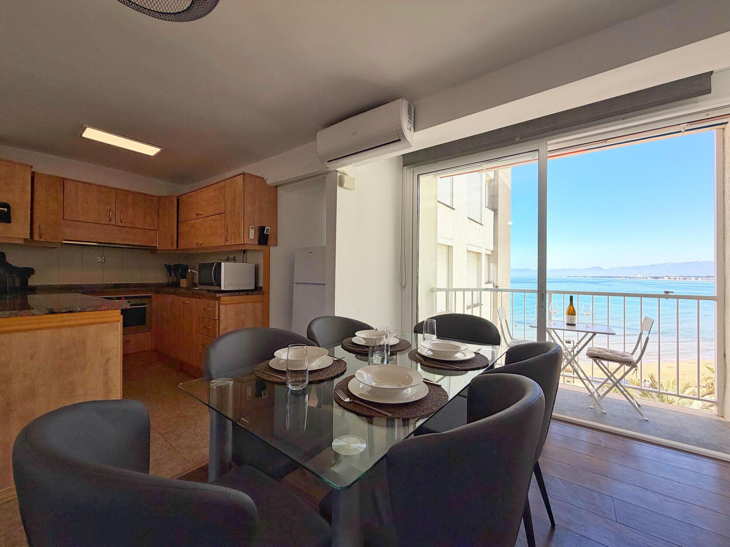 A modern kitchen with wooden cabinets, a dining area with a glass table and chairs, and a balcony with a view of the sea.