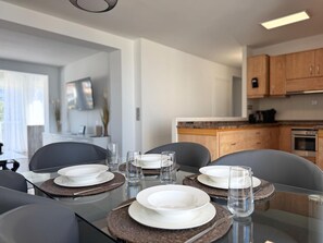 A dining table set with plates, glasses, and cutlery in a modern kitchen with wooden cabinets and a glass-top table. - Costa Dorada Apartment in Salou - Beach & Wi-Fi Included (Salou)