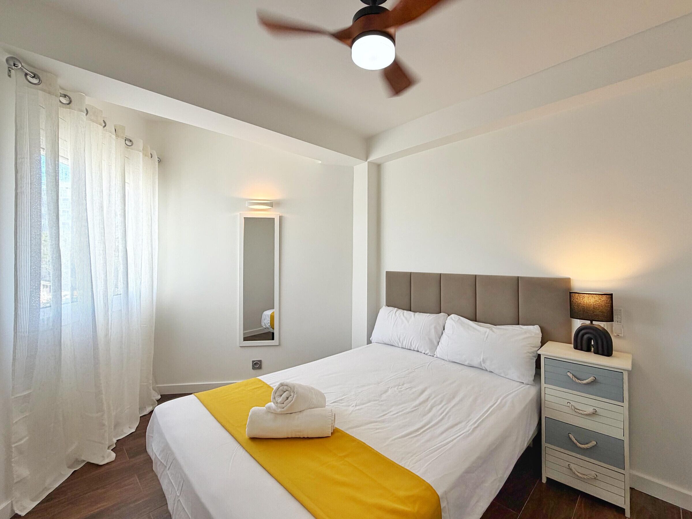 Une chambre d’hôtel avec un lit, une table de chevet avec une lampe, un miroir et un ventilateur de plafond.