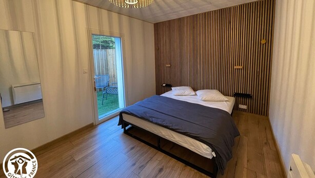 A modern bedroom with a bed, a wooden slatted wall, a ceiling light fixture, and a door leading to an outdoor area. - Family Home 700m from Beach, Spa, Pets Allowed (Saint-Hilaire-de-Riez)