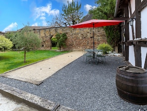 Un bâtiment en pierre avec une terrasse en gravier, un parasol rouge et une table en métal entourée de chaises.