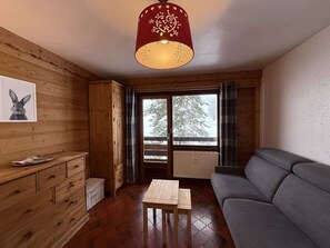 A cozy room with wooden walls, a grey sofa, a wooden table, and a window with a view of trees. - Studio for 2 Near Center Parking Included (Le Grand-Bornand)
