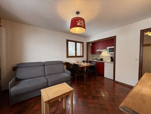 A compact living space with a grey sofa, a wooden table, a dining area with chairs, a kitchen with red cabinets, and a window with a view. - Studio for 2 Near Center Parking Included (Le Grand-Bornand)