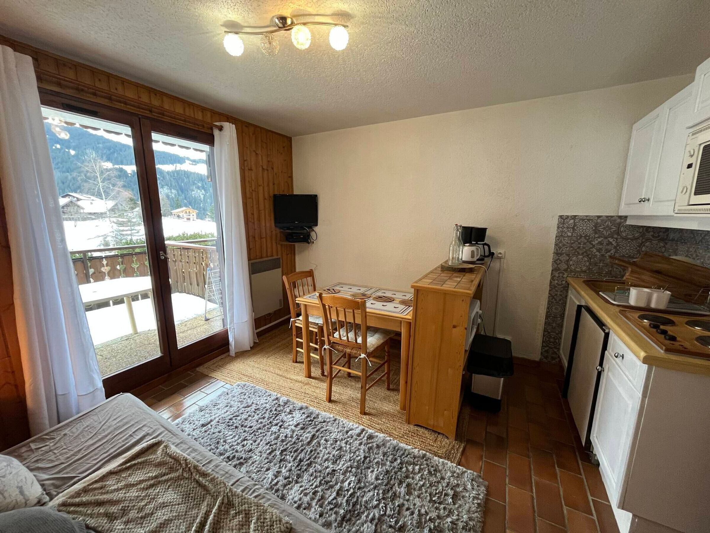 A cozy room with a bed, a small table with chairs, a kitchenette, and a sliding glass door leading to a balcony.