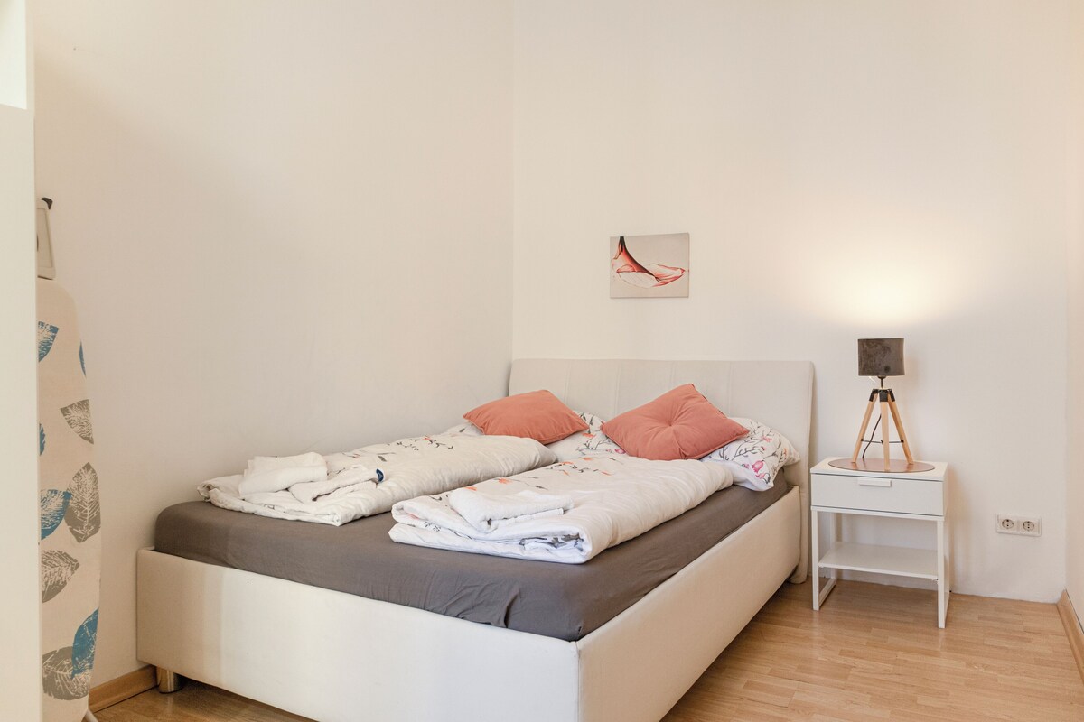 Ein ordentlich bezogenes Doppelbett mit Kissen, ein Nachttisch mit Lampe und ein an der Wand hängendes Kunstwerk.