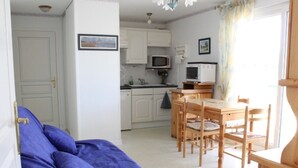 A compact living space with a sofa, a dining table with chairs, a kitchen area with cabinets and appliances, and a door leading to another room. - Apartment near sea with private parking, quick beach access (Fort-Mahon-Plage)