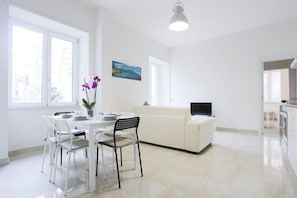 Eine moderne Küche mit weißen Geräten, ein Essbereich mit weißem Tisch und Stühlen sowie ein großes Fenster.