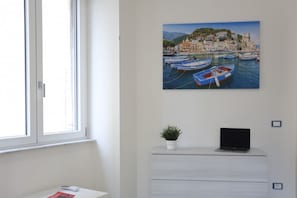 Ein Zimmer mit Fenster, Schreibtisch mit Laptop, kleiner Topfpflanze und einem an der Wand hängenden Gemälde mit Booten.