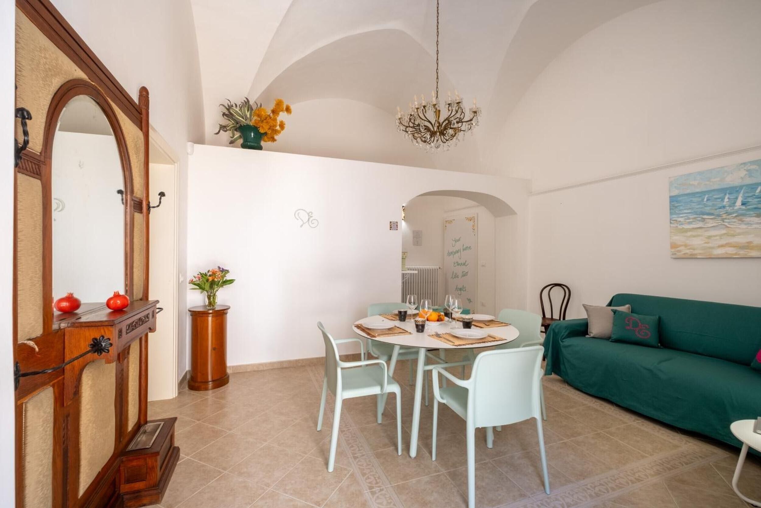 A dining area with a round table, chairs, a chandelier, a mirror, a green sofa, and a painting on the wall.