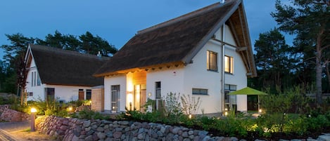 A two-story house with a stone wall in front, a thatched roof, and a well-lit entrance.