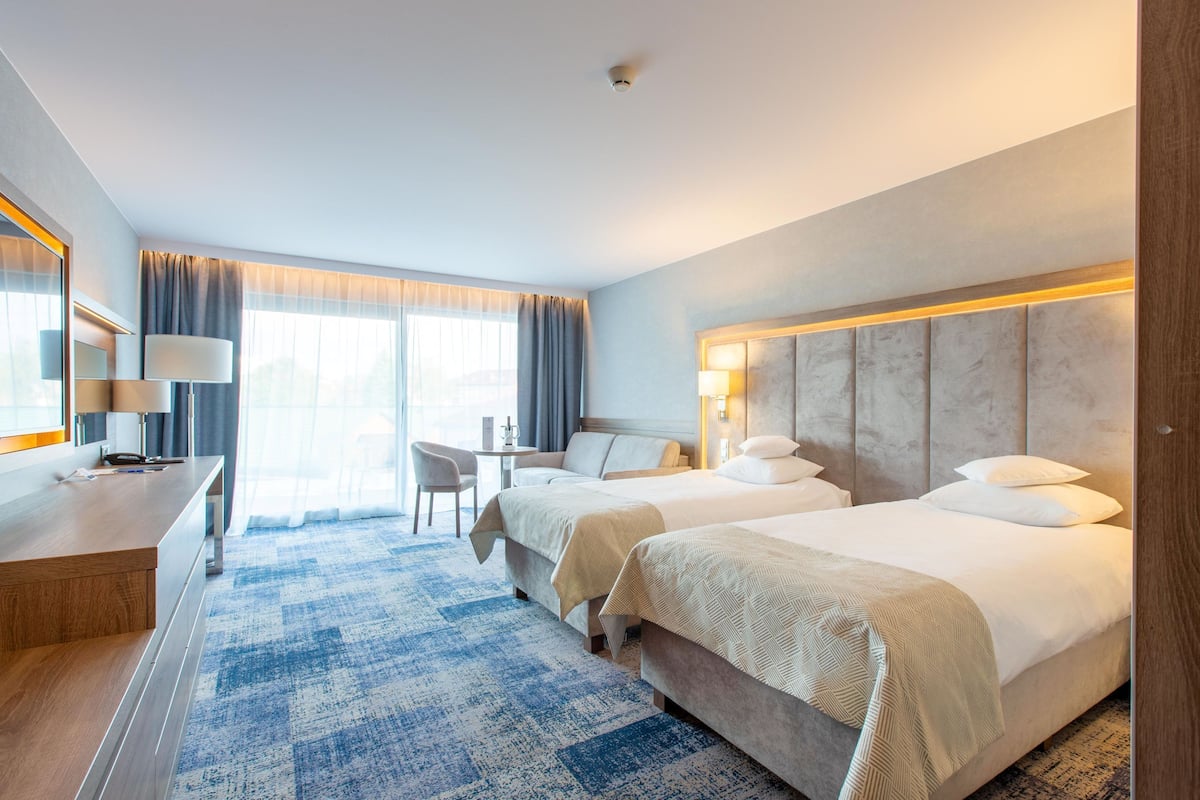 A hotel room with two beds, a desk, a chair, and a large window with curtains.
