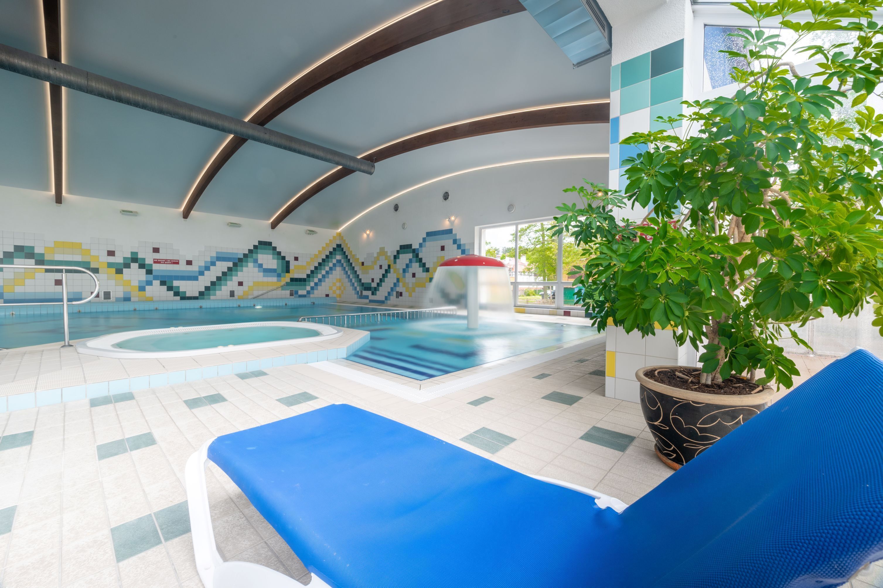 An indoor swimming pool with blue lounge chairs, a large potted plant, and a colorful mosaic wall.