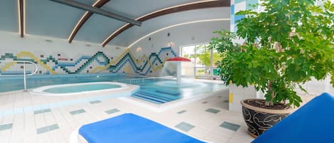An indoor swimming pool with blue lounge chairs, a large potted plant, and a colorful mosaic wall.