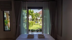 Ett hotellrum med en stor säng, vita sängkläder och utsikt över en trädgård och byggnader utanför.