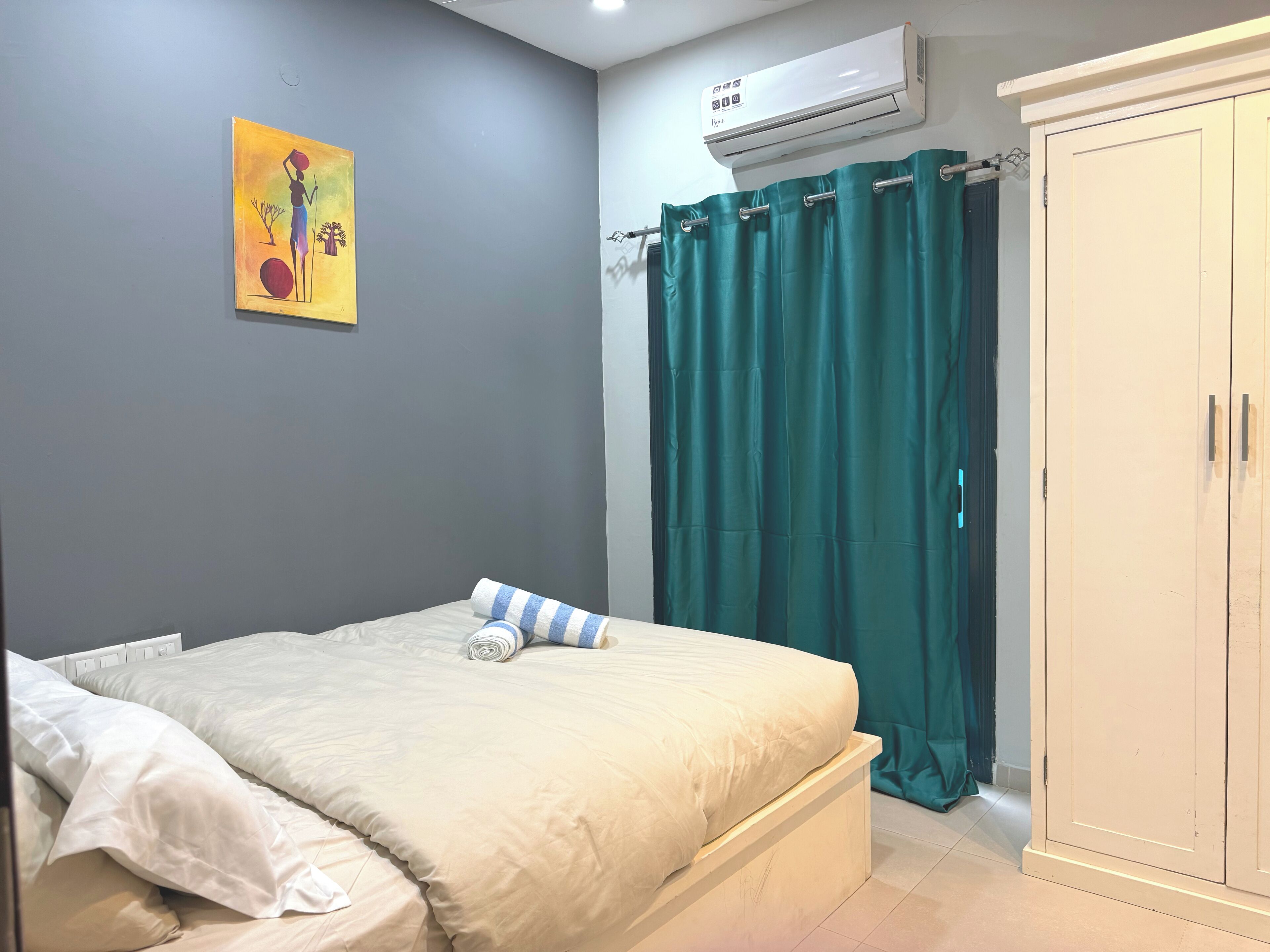 A bedroom with a bed, a painting on the wall, an air conditioner, and a wardrobe.