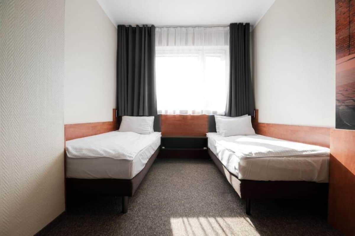 A small, compact hotel room with two single beds, a small table, and a window with curtains.