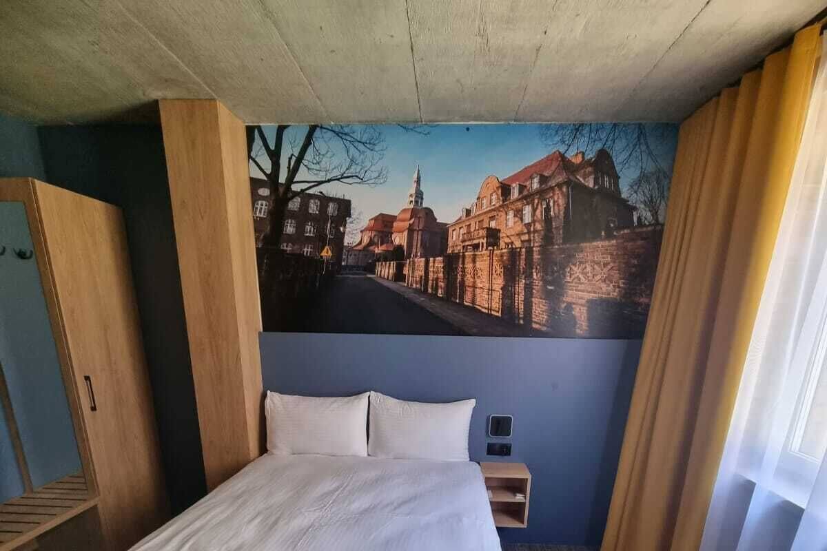 A hotel room with a large bed, a nightstand, a wooden wardrobe, and a mural of a cityscape on the wall.