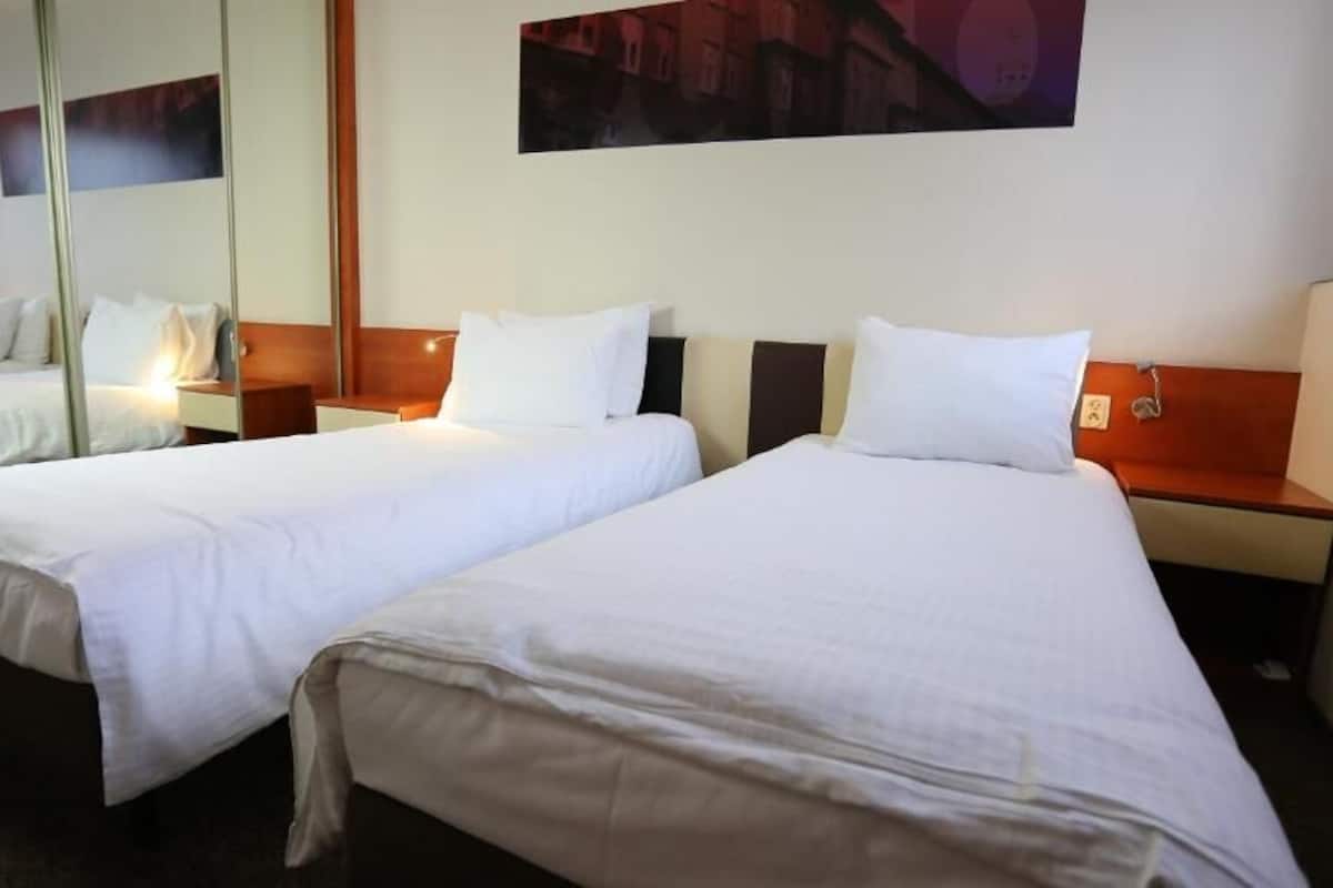 A hotel room with two single beds, each with a wooden headboard and white bedding. There are bedside tables with lamps and a large mirror on the wall.
