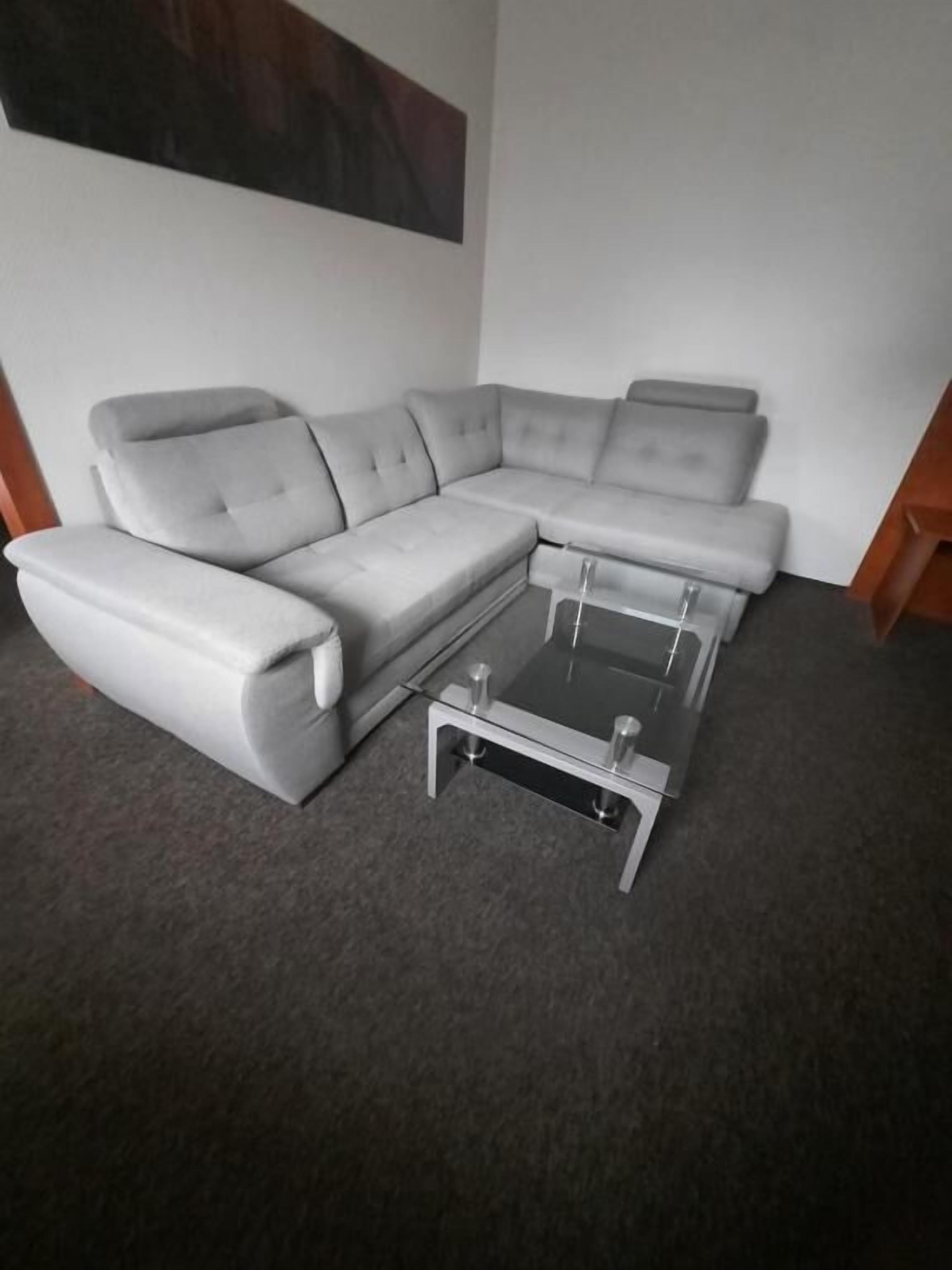 Un canapé d’angle gris, accompagné d’une table basse en verre, se trouve dans une pièce dont le sol est recouvert d’une moquette et dont le mur est uni.