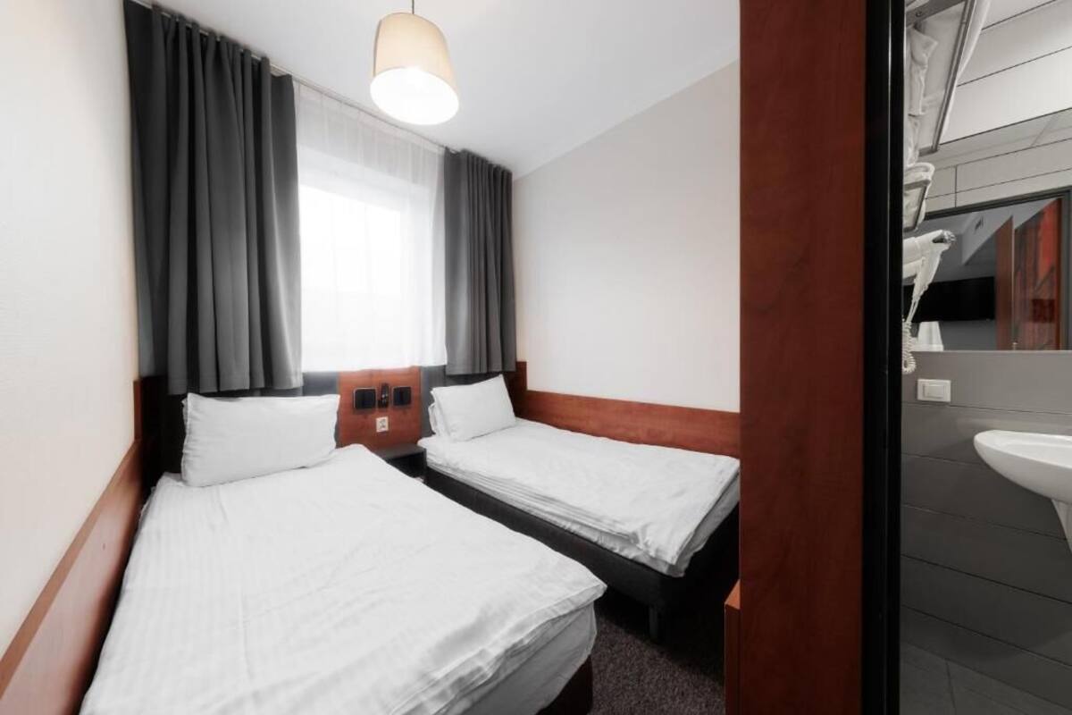 A hotel room with two single beds, a wooden headboard, a large window with curtains, a bedside table with a lamp, and a view of the bathroom.