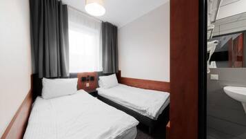 A hotel room with two single beds, a wooden headboard, a large window with curtains, a bedside table with a lamp, and a view of the bathroom.