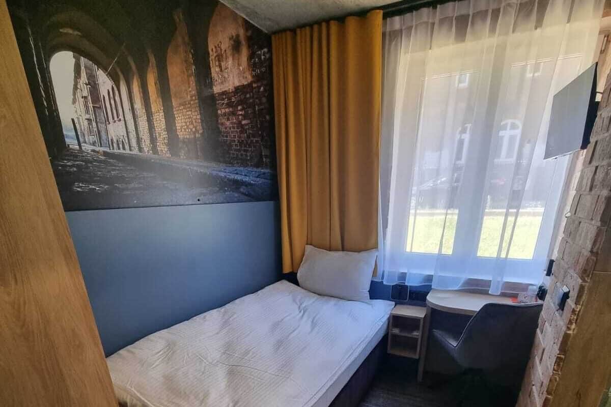 A compact room with a single bed, a small desk, and a window with curtains.