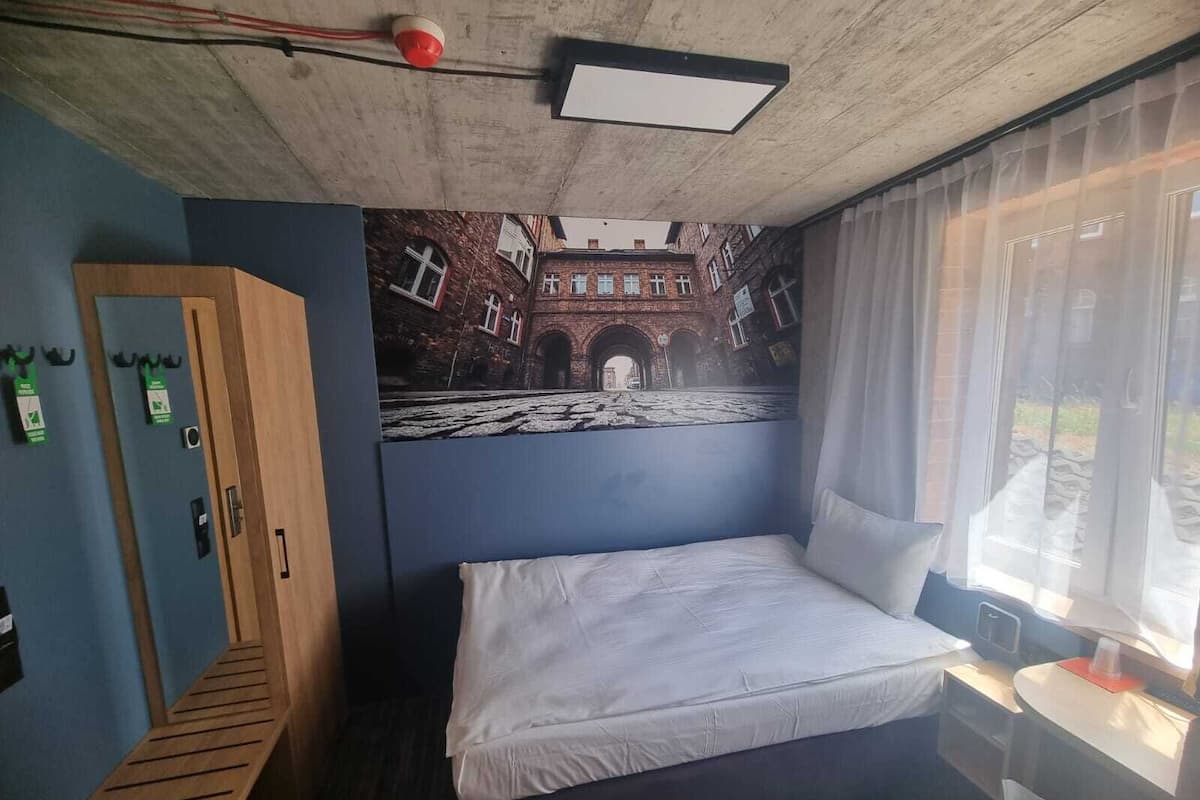 A hotel room with a bed, a wooden bench, a desk with a lamp, a window with curtains, and a mural of a building on the wall.