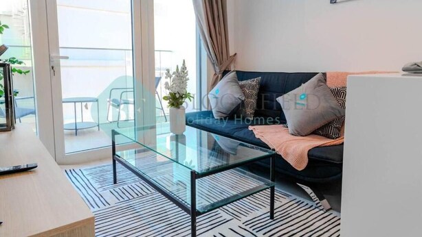 Un salon moderne avec une table basse en verre, un canapé agrémenté de coussins à motifs et une vue sur un balcon aménagé avec du mobilier d’extérieur.