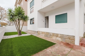 Ein modernes Gebäude mit weißer Fassade, ein kleiner Garten mit Kunstgrashalm und einer Palme.