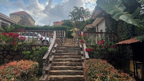 Uma escadaria de pedra leva a uma casa com telhado vermelho, rodeada por vegetação exuberante e plantas floridas.