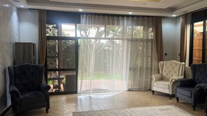 Uma sala de estar ampla com uma grande porta de vidro deslizante dando para um jardim, duas poltronas estofadas, um sofá e uma televisão sobre um móvel.