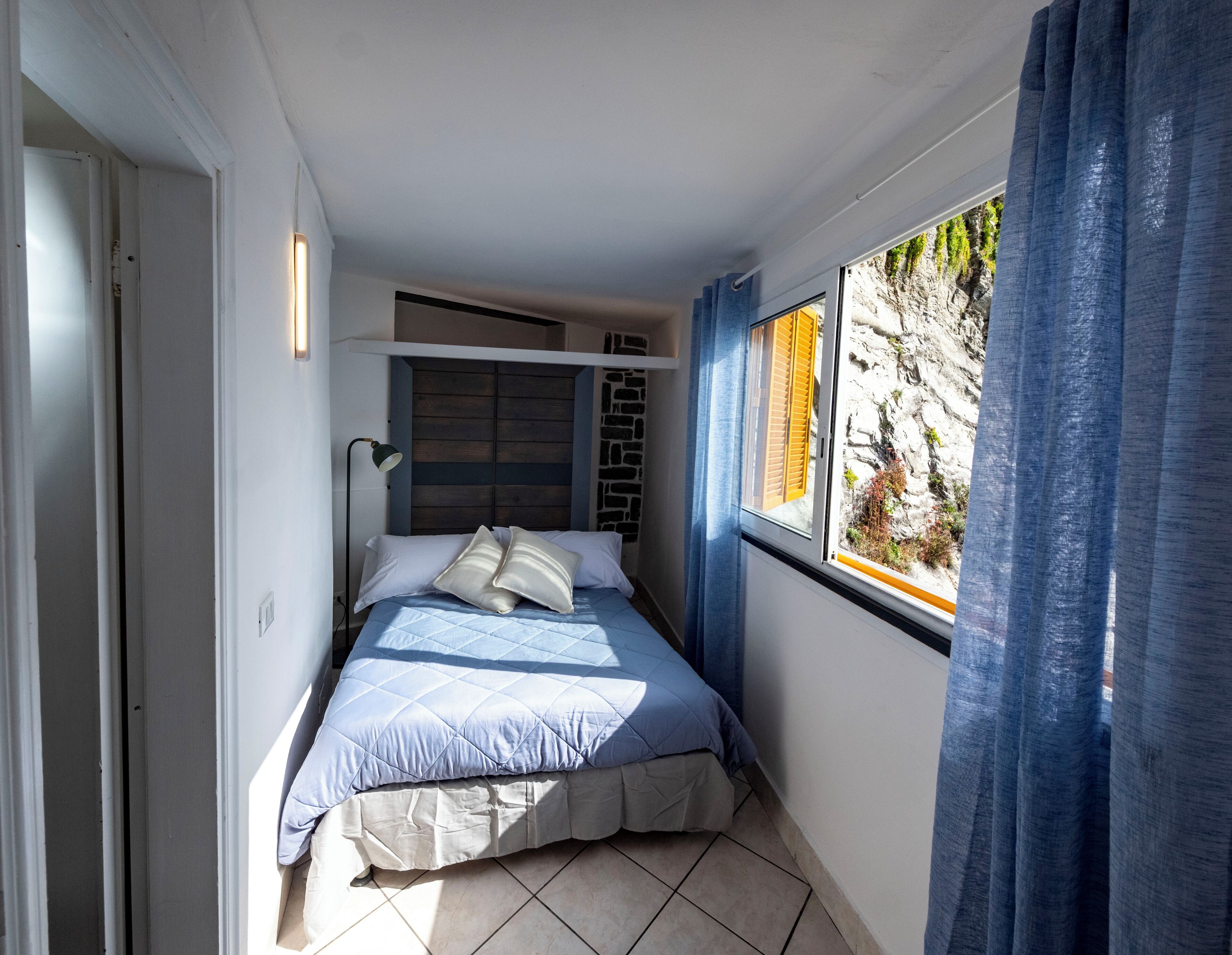 Une chambre à coucher avec un lit, une fenêtre avec des rideaux, une lampe fixée au mur et une vue sur un paysage rocheux.