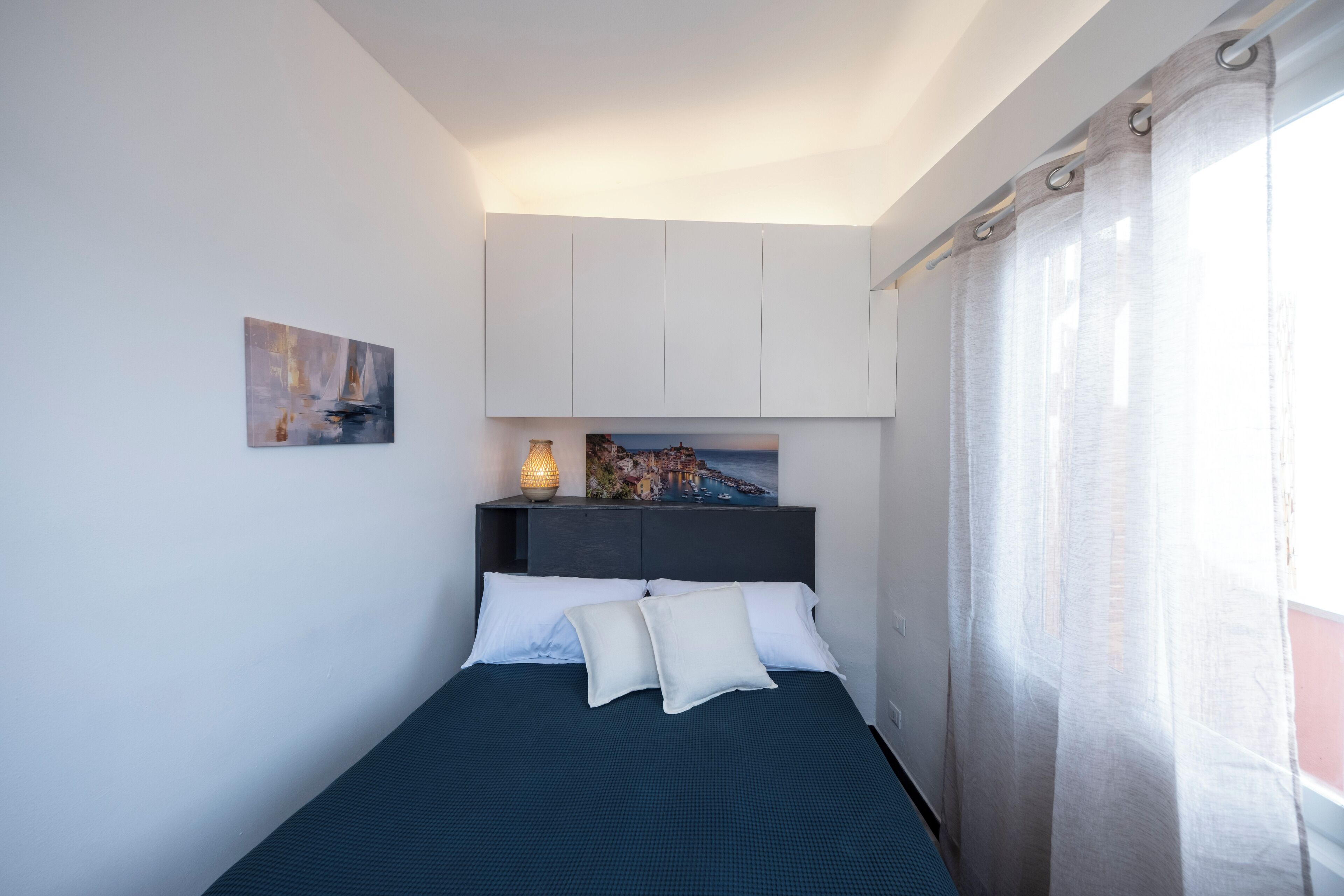 A bed with white pillows, a dark blue bedspread, a nightstand with a lamp, a painting above the bed, and a window with curtains.