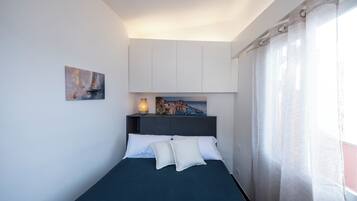 A bed with white pillows, a dark blue bedspread, a nightstand with a lamp, a painting above the bed, and a window with curtains.