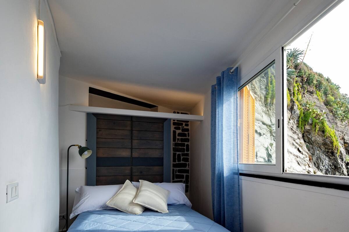 A bedroom with a bed, a nightstand, a lamp, a window with a view of a rocky landscape, and a wall with a built-in shelf.