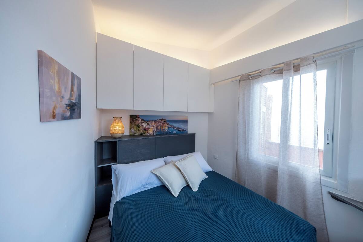 A bedroom with a bed, a nightstand, a lamp, a painting, and a window with curtains.