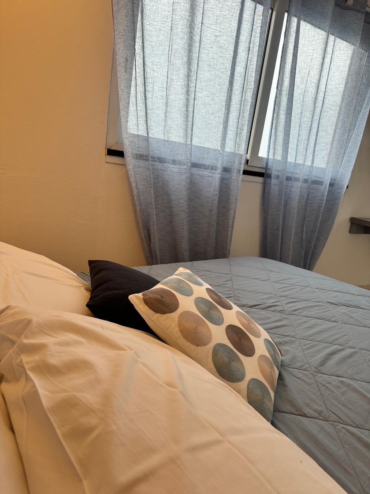 Een bed met wit en beige beddengoed, een gestreept kussen en lichte gordijnen die natuurlijk licht binnenlaten.