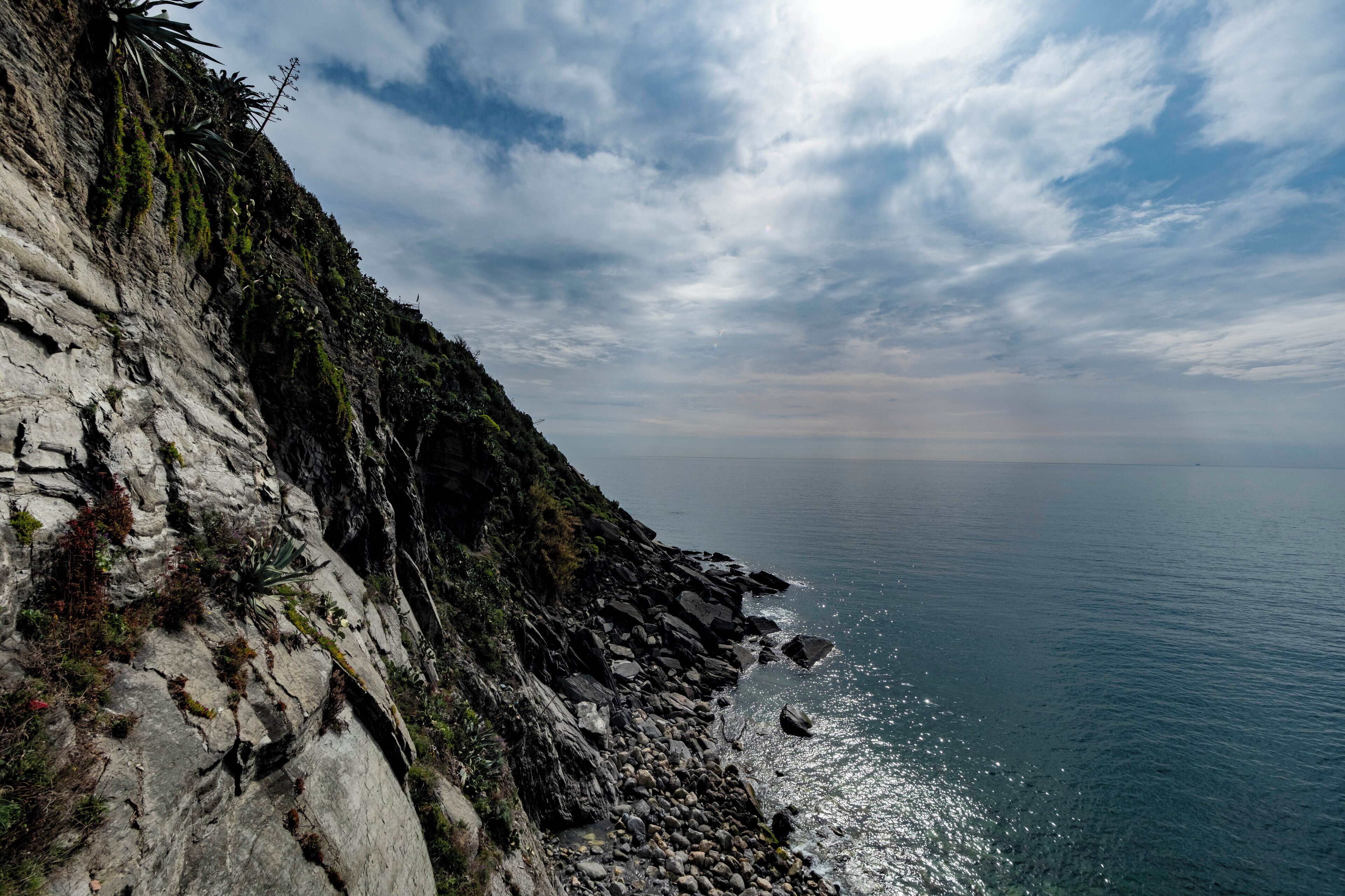 Un paysage côtier avec une falaise rocheuse, de la végétation et une mer calme sous un ciel partiellement nuageux.