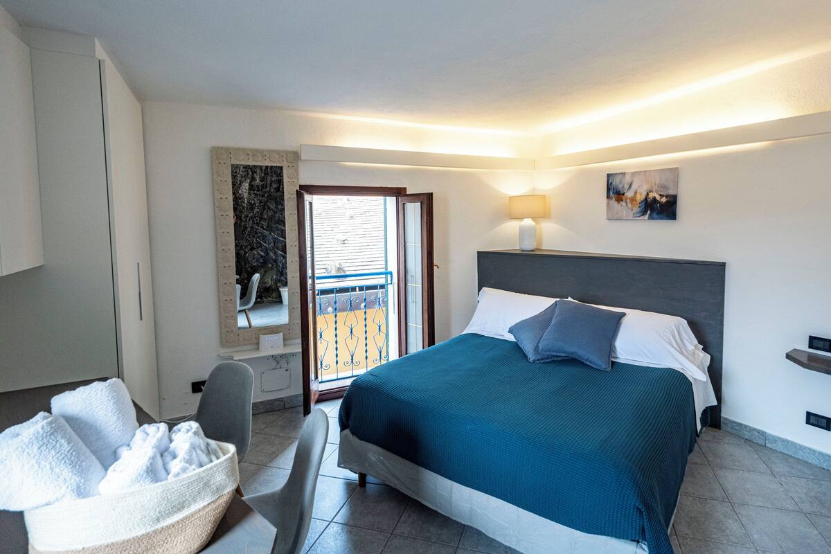 A hotel room with a bed, a chair, a desk, a lamp, and a balcony.