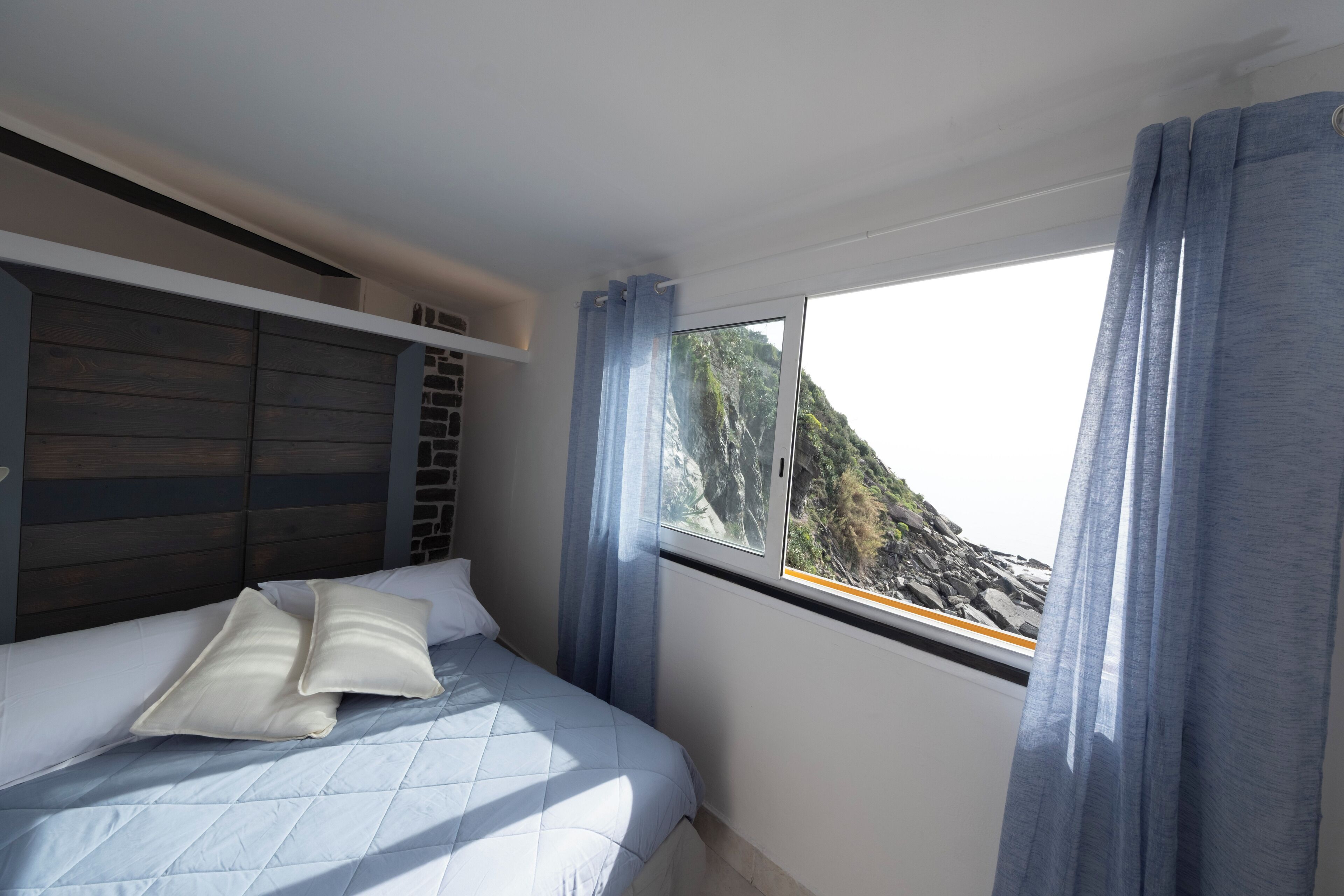 Een slaapkamer met een bed, een raam met gordijnen en uitzicht op een bergachtig landschap.
