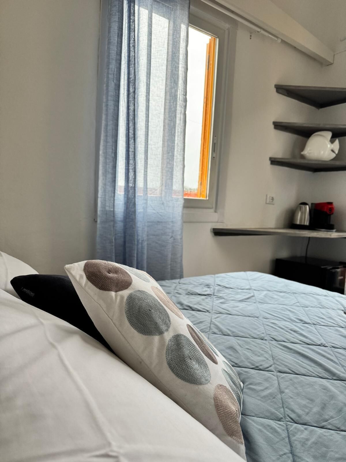 A bedroom with a bed, a window with curtains, a shelf with a kettle, and a coffee maker.