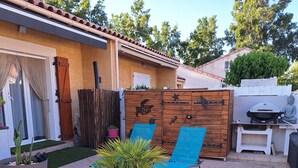 Un espace patio avec une pelouse artificielle, deux fauteuils de jardin bleus, des plantes en pot, une clôture en bois agrémentée d’éléments décoratifs et un barbecue.