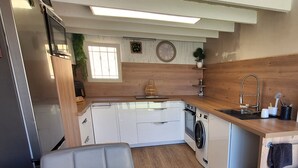 Une cuisine compacte avec des meubles blancs, un lave-linge encastré, une table à manger en bois et une chaise recouverte d’un tissu gris.