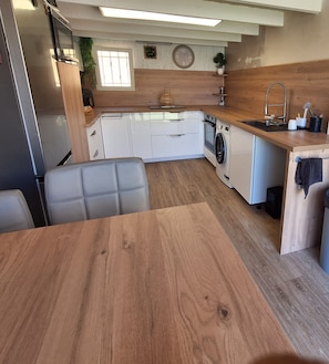 A compact kitchen with white cabinets, a built-in washing machine, a wooden dining table, and a gray upholstered chair.