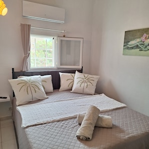A bedroom with a bed, pillows, a window, an air conditioner, and a painting on the wall.